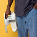 Close-up of person in jeans holding white slide sandals. Straps show a caduceus (winged staff with two snakes), 'STAYING ALIVE' in green, and names below.