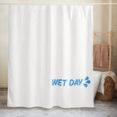 Bathroom scene showing a white shower curtain with light blue ‘WET DAY’ text and splash graphic in the lower right corner. The curtain hangs in a neutral-toned bathroom with a tub, towel rack, and woven basket visible, demonstrating real-world use.