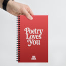 A hand holds a red spiral notebook against a white background. The cover displays the phrase 'Poetry Loves You' in large white letters and 'VIBE CHECK' in smaller text near the bottom.