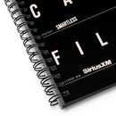 Detailed close-up of the bottom left corner of the black spiral notebook. The black metal coil binding is visible, along with white block letters spelling ‘FILES’, and the white logos ‘SmartLess’ and ‘SiriusXM’ in smaller print.