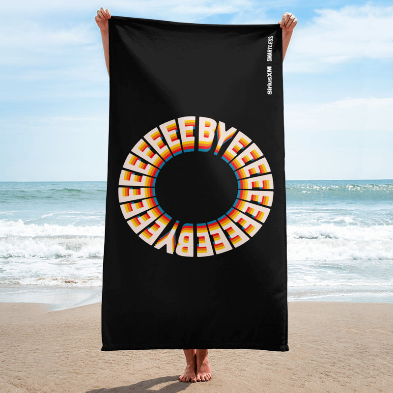 A person holds a black towel vertically on the beach, showing a large circular rainbow-styled 'BYEEEEEEE' design centered on the towel. The 'SiriusXM' and 'SMARTLESS' logos appear in white along the upper right edge.