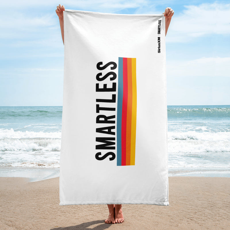 A person stands on the beach holding a vertically oriented white towel featuring the word 'SMARTLESS' in bold black letters next to four vertical stripes in blue, red, orange, and yellow. The 'SiriusXM' and 'SMARTLESS' logos are printed in black near the top right edge.