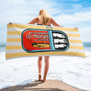 Woman standing on the beach holding a towel horizontally behind her; the towel features a sardine can illustration with the text 'Summer S’mores with Conan & the Chill Chums' on yellow and white stripes.