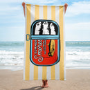 Person holding a vertically oriented towel on the beach; the towel shows a sardine can design and the same 'Summer S’mores with Conan & the Chill Chums' graphic with yellow and white stripes.