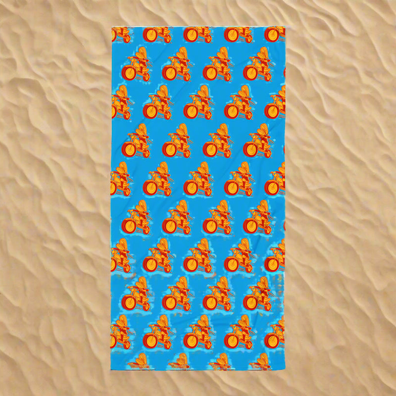 A full view of a bright blue towel laid flat on sand, covered with a repeating pattern of the Comedy Bang Bang motormouth logo character—a red and yellow motorcyclist with chattering teeth for a head.