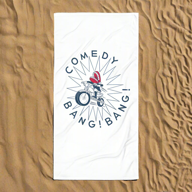 A white towel laid flat on textured sand, displaying the 'Comedy Bang! Bang!' logo with chattering teeth riding a motorcycle at the center, surrounded by a spiked starburst and bold navy text.