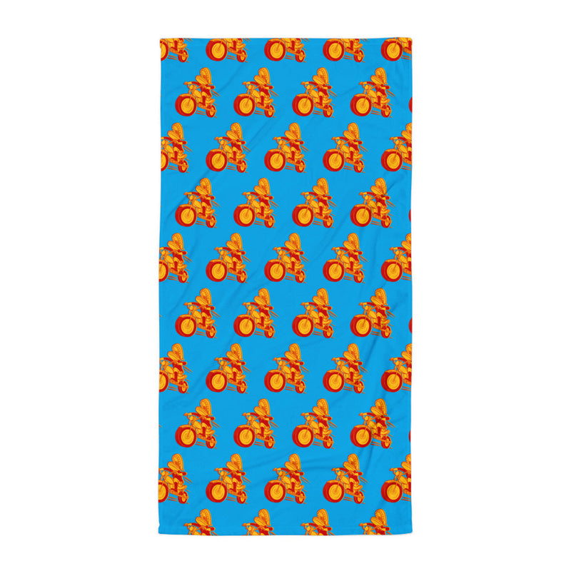 A bright blue towel laid flat on a white surface, covered with a repeating pattern of the Comedy Bang Bang motormouth logo character—a red and yellow motorcyclist with chattering teeth for a head.