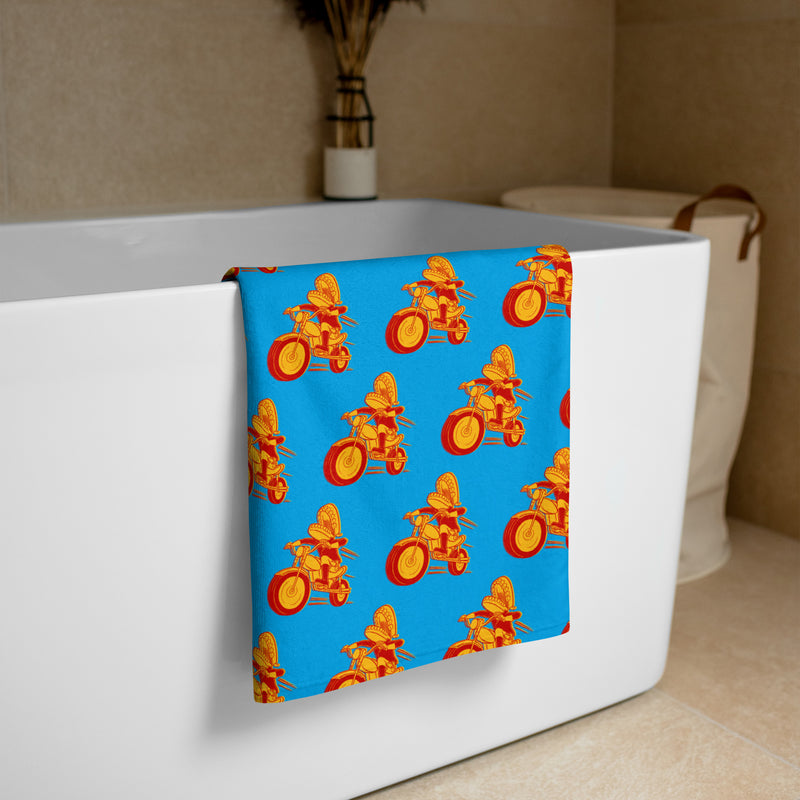 A bright blue towel featuring a repeating pattern of a red and yellow chattering teeth motorcyclist character riding a motorcycle, draped over the side of a white bathtub in a modern bathroom.