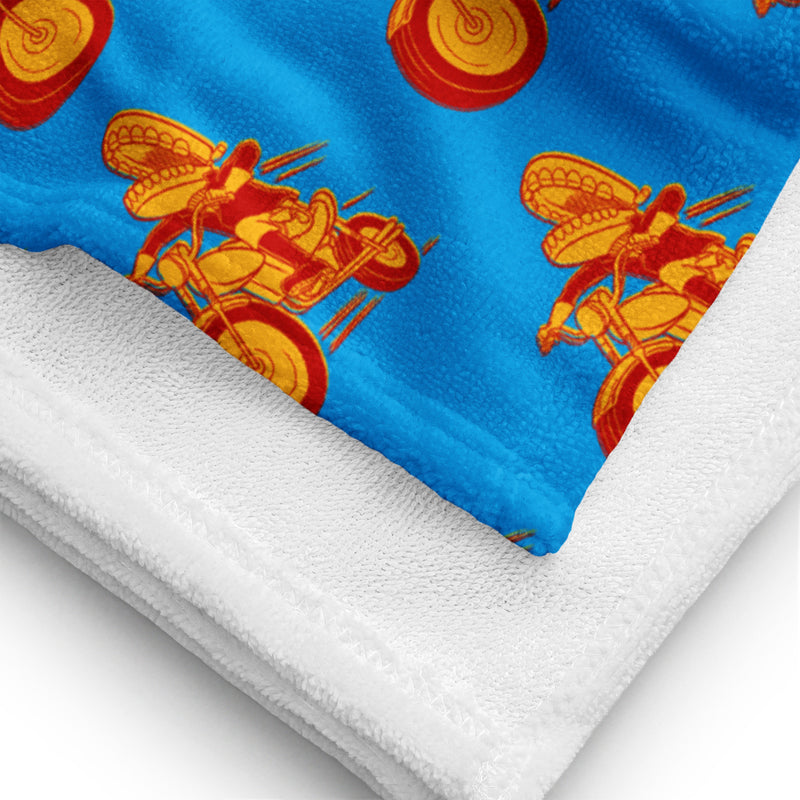 A close-up view of the blue towel showing the detailed fabric texture and stitching, along with a clear look at the Comedy Bang Bang logo pattern of a motorcyclist with a chattering teeth toy for a head in red and yellow tones.