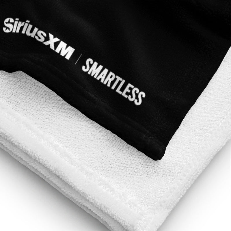 A close-up view of the corner of a black towel with white underside, featuring the 'SiriusXM' and 'SMARTLESS' logos printed in white near the edge stitching.