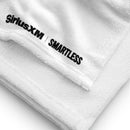 A close-up of a white towel corner, showing the stitched edge and the 'SiriusXM' and 'SMARTLESS' logos printed in black near the hem on the soft terry fabric.