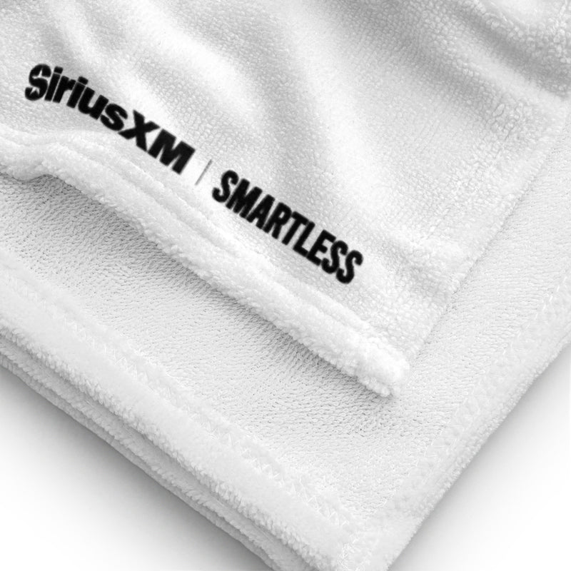 A close-up of a white towel corner, showing the stitched edge and the 'SiriusXM' and 'SMARTLESS' logos printed in black near the hem on the soft terry fabric.