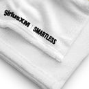 A close-up of the corner of a white towel showing soft terry texture and the 'SiriusXM' and 'SMARTLESS' logos printed in black near the edge seam.
