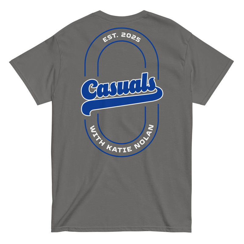Charcoal short-sleeve t-shirt laid flat, showing the large blue and white back design with 'Casuals' in bold in the center, 'EST. 2025' above, and 'WITH KATIE NOLAN' curved below.