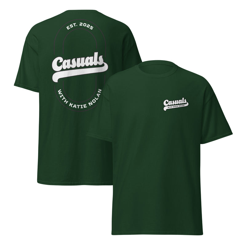 Forest green short-sleeve t-shirt shown from both angles. Front: small white logo on the left chest reading 'Casuals' with 'WITH KATIE NOLAN' below. Back: large oval outline with 'Casuals' in bold white text, 'EST. 2025' above, and 'WITH KATIE NOLAN' curved below.