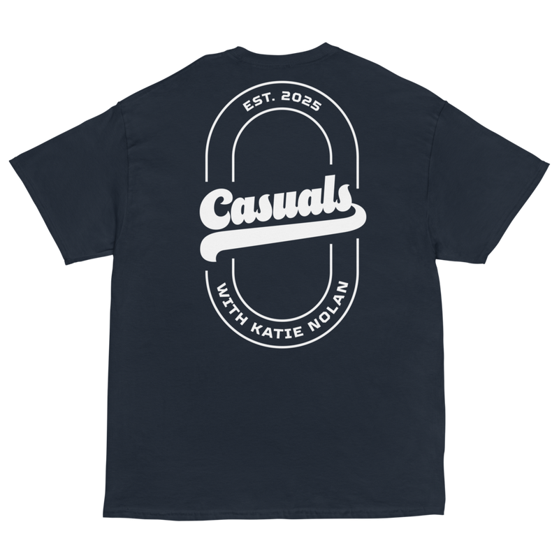 Navy short-sleeve t-shirt laid flat, featuring a large white oval outline on the back with bold text 'Casuals' in the center, 'EST. 2025' above, and curved text 'WITH KATIE NOLAN' below.