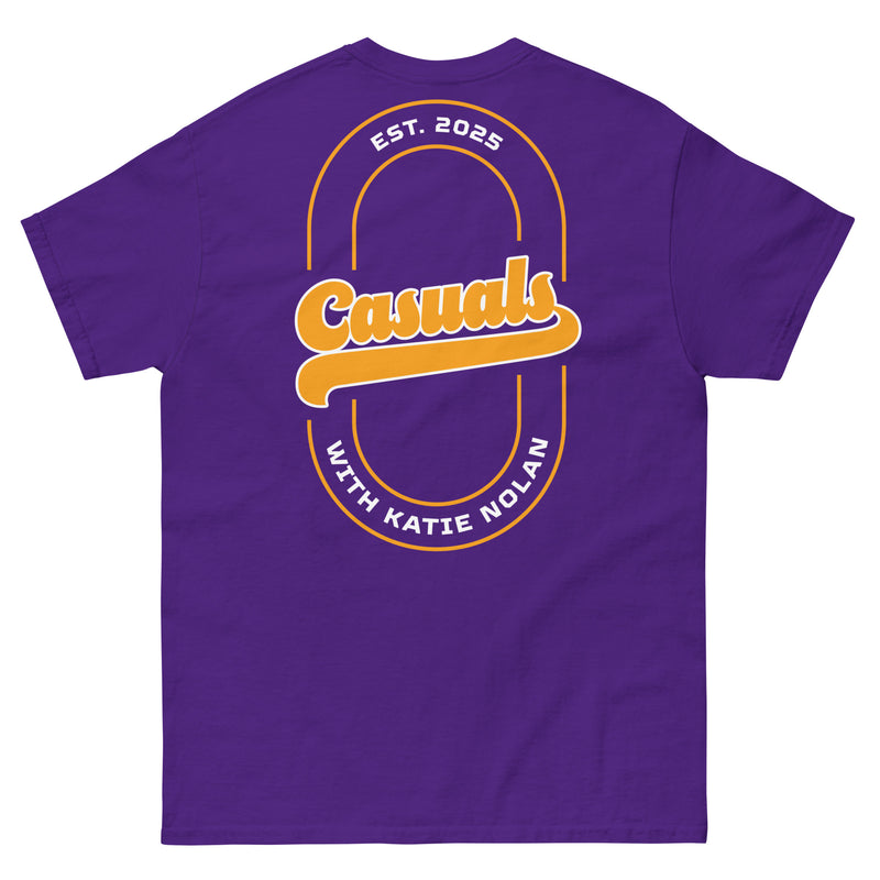 Purple short-sleeve t-shirt laid flat, showing the back design: a large gold and white oval outline with 'Casuals' in bold gold letters, 'EST. 2025' above in white, and 'WITH KATIE NOLAN' curved below in white.