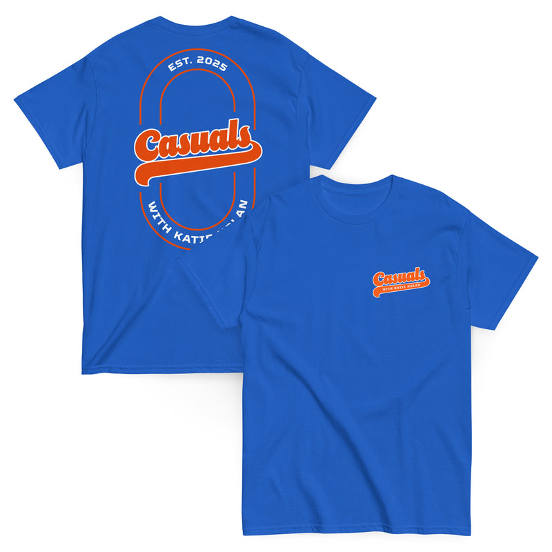 Two royal blue short-sleeve T-shirts laid flat, one showing the front and one showing the back. The front displays a small orange and white 'Casuals WITH KATIE NOLAN' logo on the left chest, and the back features a large orange 'Casuals' design with the text 'EST. 2025' and 'WITH KATIE NOLAN' arranged in a circular layout within a double-line oval.