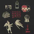 Close-up of the back graphic on a black heavyweight T-shirt. Centered is a collage-style layout featuring vintage-inspired illustrations: a pair of overlapping skulls in red and black, a stack of old books, a crystal ball, a raven perched on a branch, a small horned devil figure, a lit candle in red, a decorative gothic window frame, and a cropped image of wide, fearful eyes. In the center, the word ‘MORBID’ appears stacked three times in red capital letters.