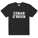 Front view of a black unisex garment-dyed heavyweight T-shirt featuring bold white block lettering centered on the chest that reads ‘CONAN O’BRIEN’. The text is stacked in two lines in a varsity-style font, with ‘CONAN’ above ‘O’BRIEN’. The shirt has a classic crew neck, short sleeves, and a relaxed fit.