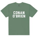 Front view of a light green unisex garment-dyed heavyweight T-shirt featuring large white varsity-style block lettering centered on the chest that reads ‘CONAN’ on the top line and ‘O’BRIEN’ below. The bold stacked text contrasts cleanly against the muted green fabric. The shirt has a crew neck and short sleeves with a relaxed fit.