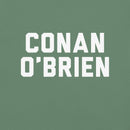 Close-up of the centered chest graphic on a light green T-shirt showing thick white varsity-style block letters spelling ‘CONAN O’BRIEN’ stacked in two lines. The crisp white lettering stands out sharply against the soft sage-green cotton fabric, with clean edges and evenly spaced alignment clearly visible.