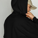 Close-up side view of a person wearing a beige cap and a black hoodie with the hood up, holding the edge of the hood near the face; no design is visible.