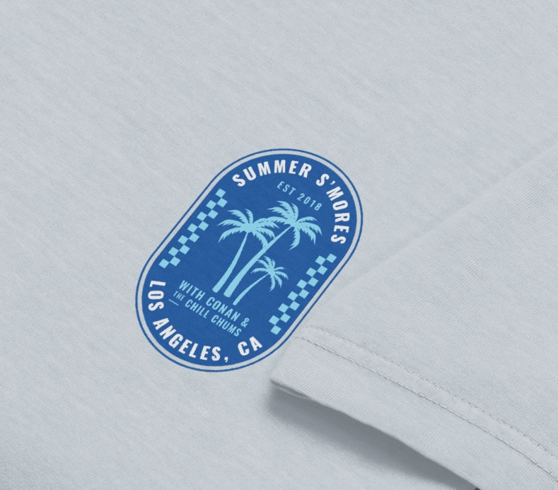 A detailed view of the printed chest logo showing palm trees and the text ‘Summer S’mores with Conan & the Chill Chums, Los Angeles, CA’ on a light blue garment.