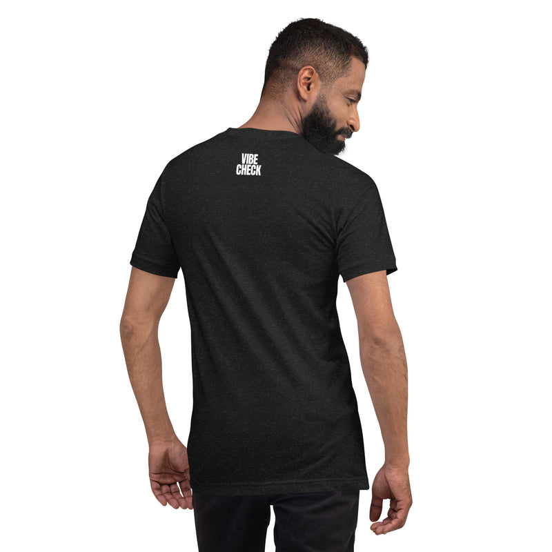 A man wearing a black heather t-shirt is shown from behind, turning slightly to his left. The back of the shirt features the phrase 'VIBE CHECK' in small white capital letters printed just below the neckline. The man has a short beard and is wearing black pants, standing against a plain white background.