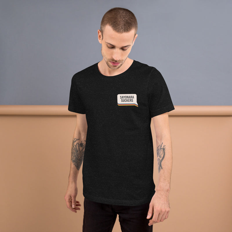 A man with arm tattoos wearing a black heather T-shirt featuring a chest graphic that reads 'SAYONARA SUCKERS' with a small rainbow stripe underneath.