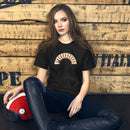 A woman sitting on the floor against a wooden backdrop, wearing a black heather T-shirt with the arched 'BYEEEEEEEE' design printed across the front.