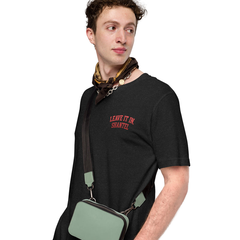 A man looking to the side while wearing a black heather T-shirt with red text 'LEAVE IT IN, SHANTEL' on the left chest, accessorized with a brown patterned scarf and a mint green crossbody bag.