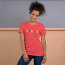 A smiling woman wearing a heather red T-shirt with the word 'BYEEEEEEEE' printed across the chest in bold, colorful letters. She is standing casually with one hand in on her thigh.