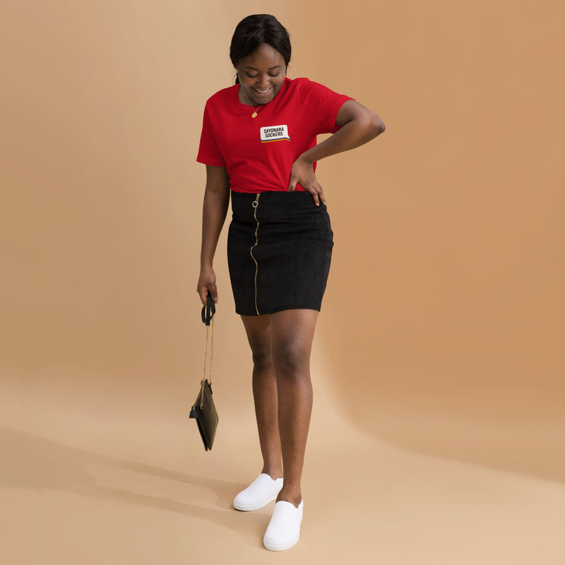 A smiling woman wearing a red T-shirt with the phrase 'SAYONARA SUCKERS' in a speech-bubble graphic on the front, paired with a black zip-up skirt and white slip-on shoes.
