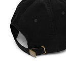 Close-up of the back of a black corduroy cap showing the ribbed fabric texture and stitched eyelets. The adjustable strap passes through a brass-tone metal slider buckle with a rectangular plate and curved clasp. The back opening forms a half-moon shape beneath the crown, highlighting the cap’s structured panels and thick corduroy weave.