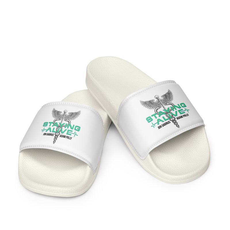 A pair of white slides shown on a plain surface with a green and gray 'STAYING ALIVE' logo and a caduceus symbol, a winged staff entwined with two snakes, printed across the top straps.