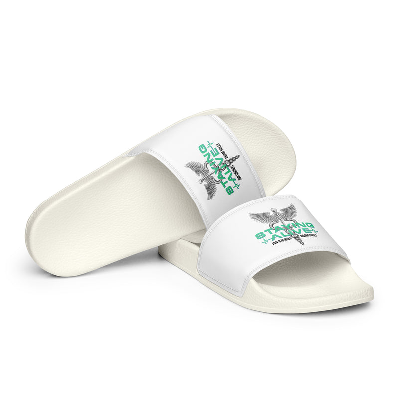 A pair of white slides placed diagonally on a white background, both featuring a green and gray 'STAYING ALIVE' graphic and a caduceus symbol, a winged staff wrapped with two snakes, printed across the foot straps.