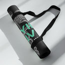 A rolled-up black yoga mat secured with black elastic straps and a carrying handle rests on a sunlit floor. The visible section of the mat features close-up details of a caduceus — the classic medical symbol composed of a winged staff and two coiled serpents — along with partially obscured teal text reading 'STAYING ALIVE' and part of the names 'JON GABRUS' and 'ADAM PALLY' printed in white.