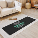A black yoga mat lies on a beige rug in a cozy living room. The mat features a gray caduceus — a winged staff with two coiled snakes, symbolizing medicine and healing — along with teal text reading 'STAYING ALIVE', flanked by heartbeat lines, and the names 'JON GABRUS' and 'ADAM PALLY' in white below.
