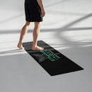 A person in black athletic shorts steps barefoot onto a black yoga mat in a bright studio. The mat displays a gray caduceus, a symbol of healthcare featuring two snakes and wings, with teal text 'STAYING ALIVE' and white text 'JON GABRUS' and 'ADAM PALLY'.